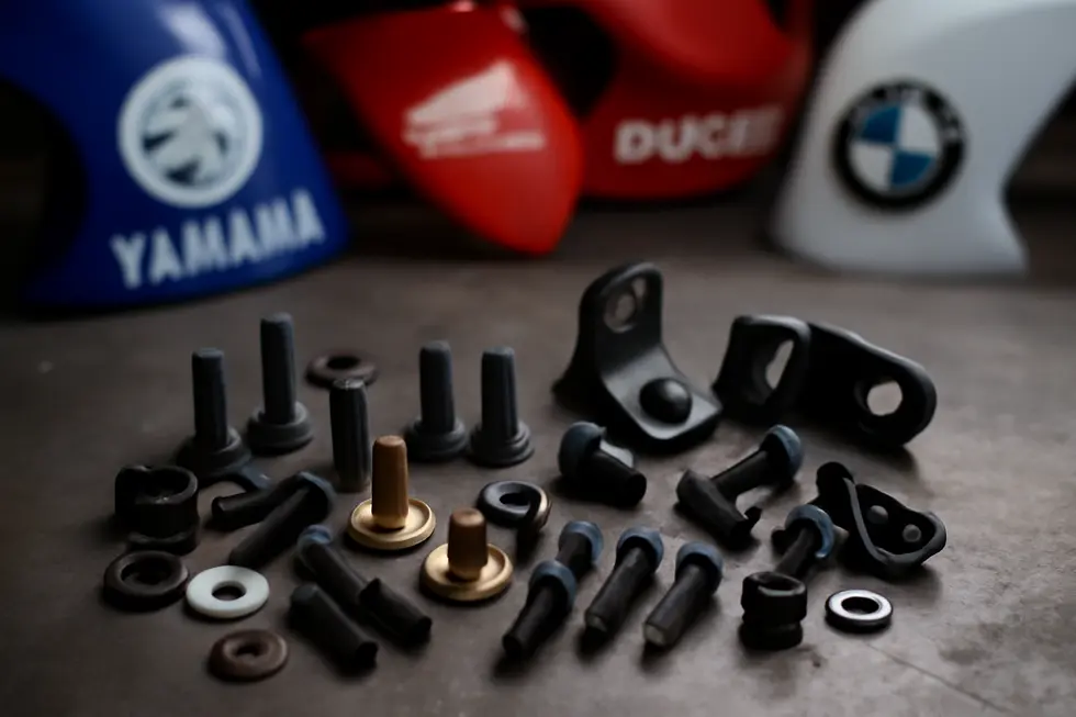 Various motorcycle fairing bolts, screws, and mounting brackets arranged on a workbench with motorcycles and brand logos softly visible behind.